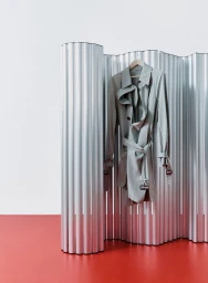 A grey trench coat hanging on a wooden hanger against a corrugated metallic screen and a red floor.