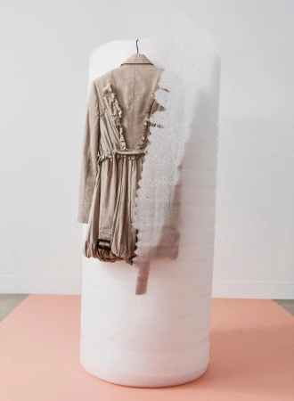 Beige trench coat hanging on a vertical white cylindrical architectural form against a plain white wall and peach floor.