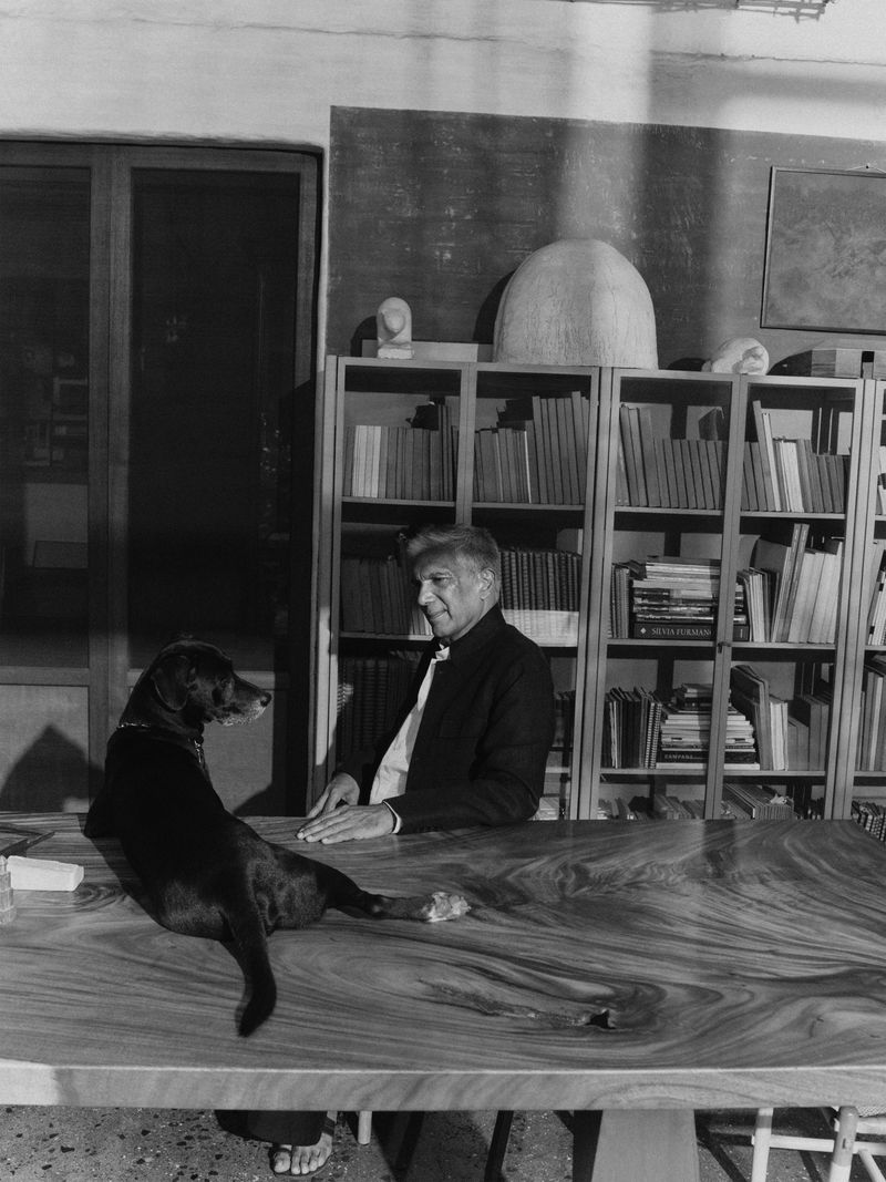 Monochrome portrait of an older man seated at a large wooden table with a black dog resting on it in a book-lined room