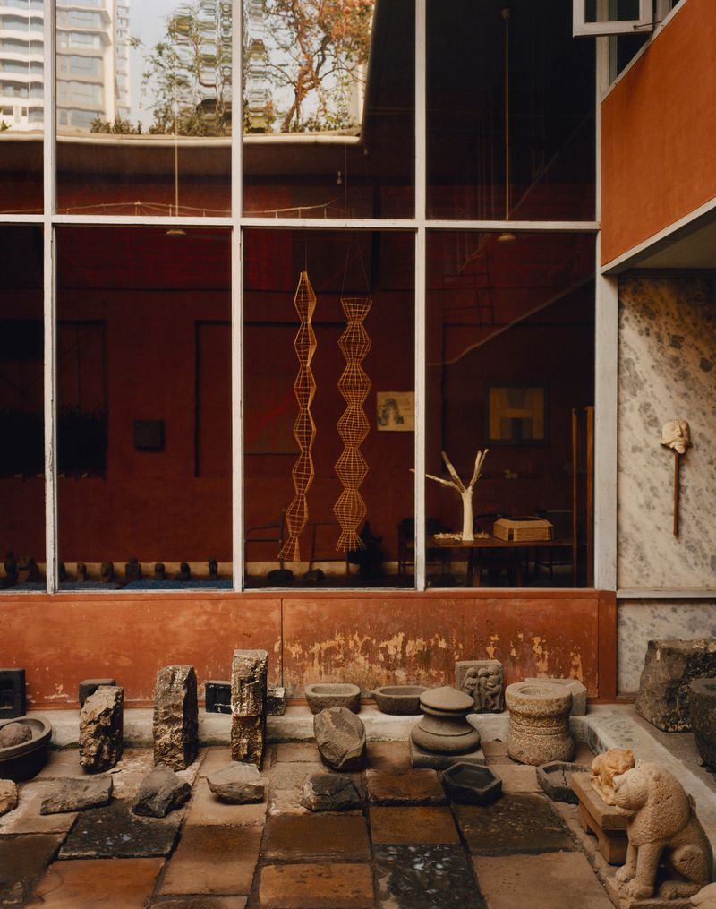 Architectural shot of an outdoor courtyard displaying a collection of stone objects and sculptures, viewed through a window with a red interior.