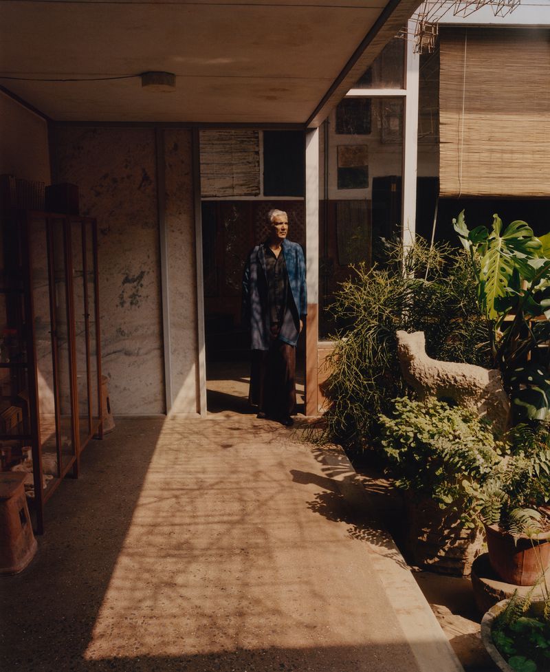 An elderly person standing in a sunlit doorway of a porch with potted plants and architectural details for Justsmile Magazine