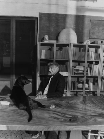 Monochrome portrait of an older man seated at a large wooden table with a black dog resting on it in a book-lined room