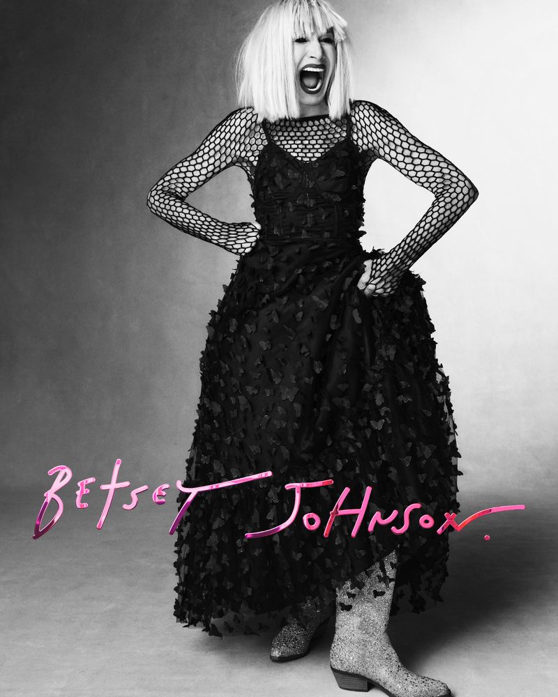 A woman in a black dress with a fishnet top and sparkly boots, standing with hands on hips and screaming, on a grey studio background.