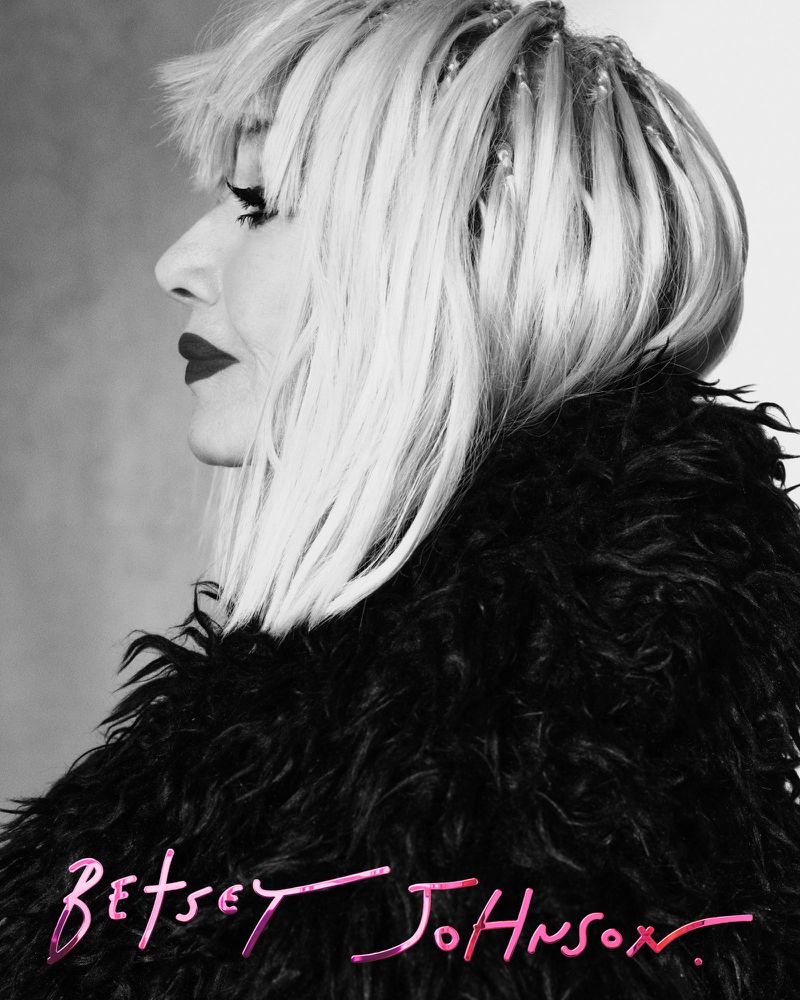 Black and white side-profile shot of an adult female with platinum-blonde hair, dark lipstick, and a black, feathery garment. 