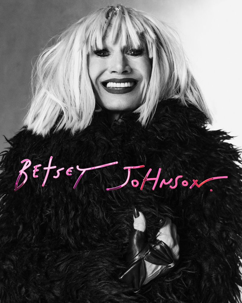 Monochrome portrait of a smiling adult woman wearing a shaggy black coat, light hair, and vibrant red lipstick against a soft gray backdrop.
