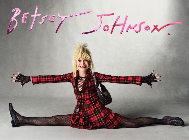 Adult woman in a red plaid dress and black fishnets sitting with her legs spread on a grey background beneath the words Betsey Johnson.