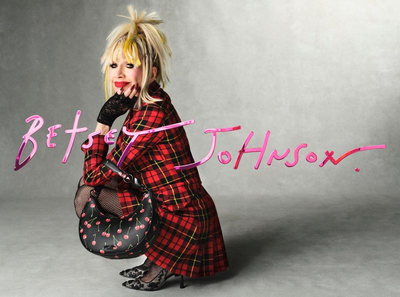 Fashion portrait of a blonde woman with red plaid coat and cherry handbag squatting in front of a light grey background.