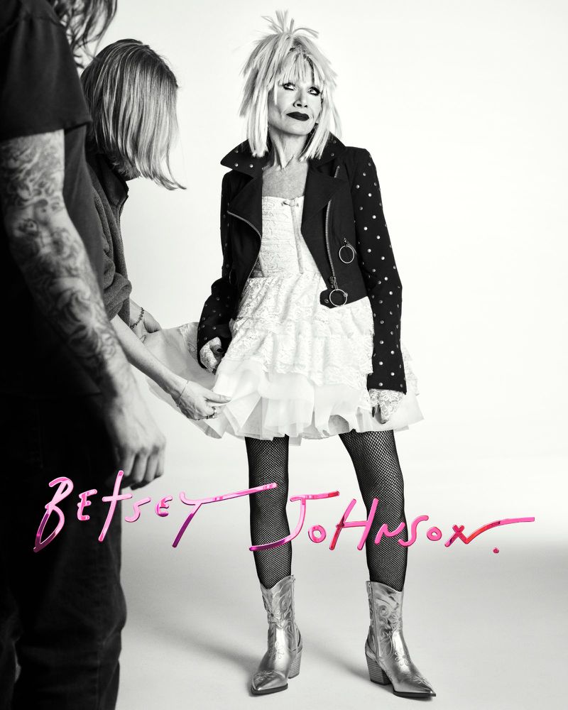 A fashion shot featuring a model in a white dress and black jacket, styled with blonde hair, and metallic cowboy boots, flanked by two people.