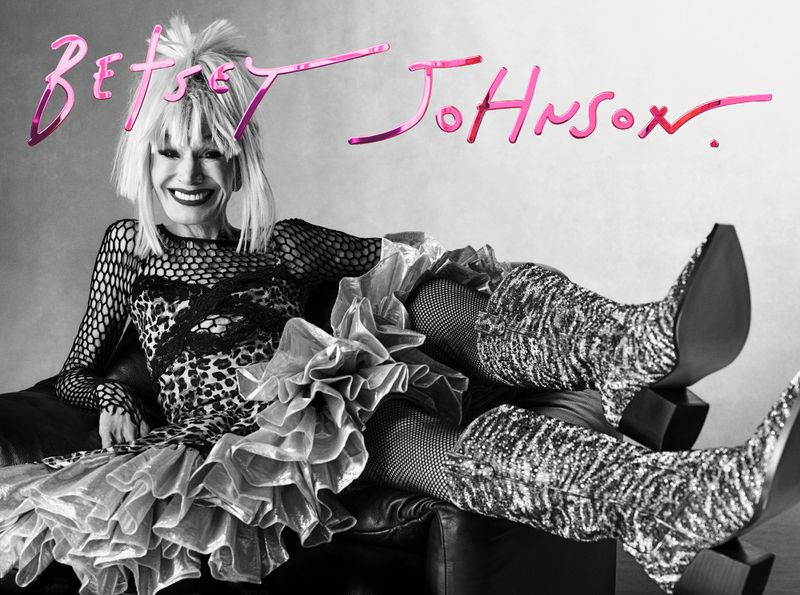 Mature woman in Betsey Johnson apparel smiles while posing on a black leather couch, with the Betsey Johnson logo in pink above.