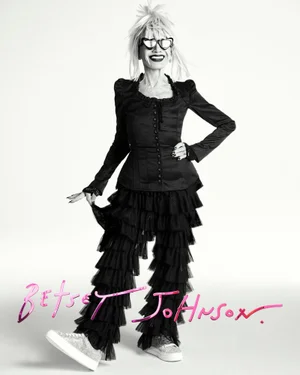 Elderly woman wearing cat-eye sunglasses and a black ruffled outfit against a white background