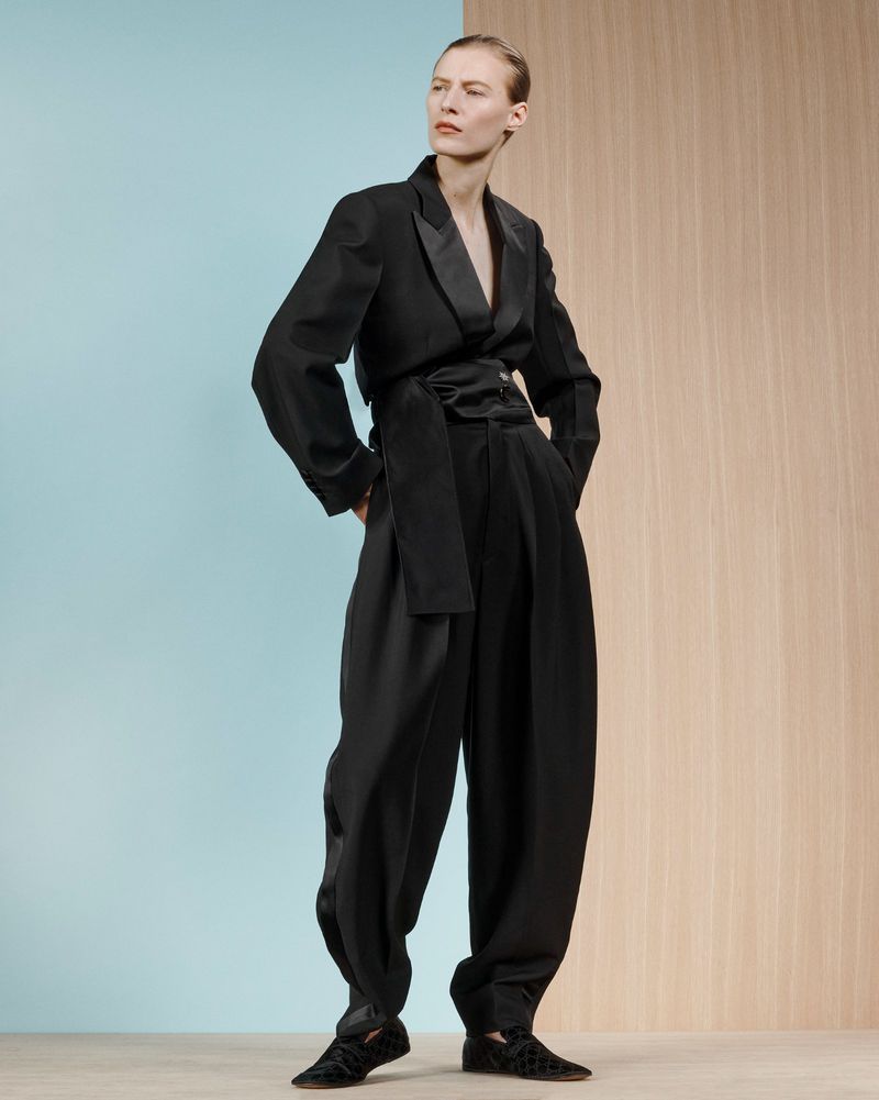 Elegant woman in a black suit with a wrap belt posing in a studio with a blue and wood-tone background. Fashion editorial style.