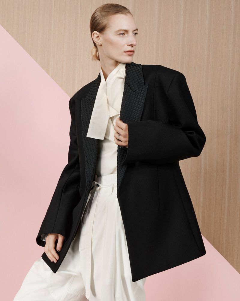 Model Julia Nobis wearing an oversized black blazer over a white blouse against a geometric pink and wood-textured backdrop.