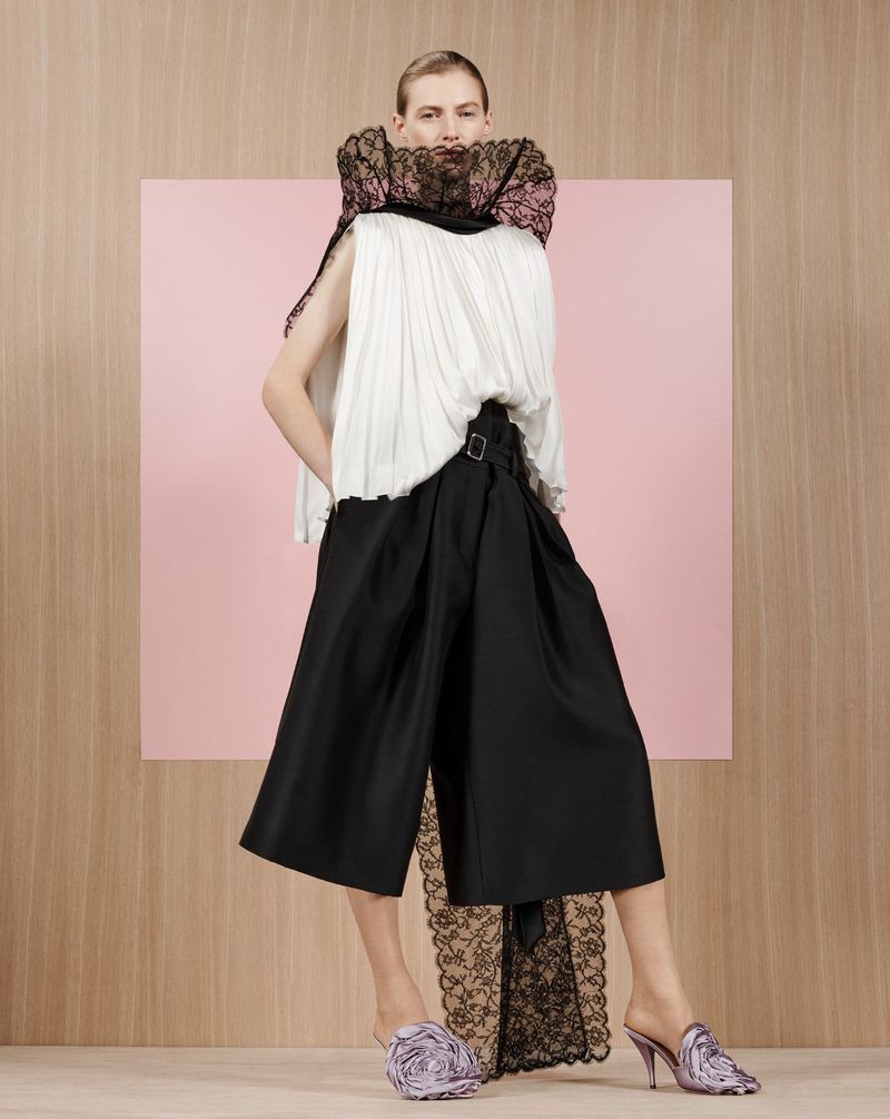 Model Julia Nobis wearing a white top, black trousers, and a black lace ruff collar against a wood-textured and pink square background.