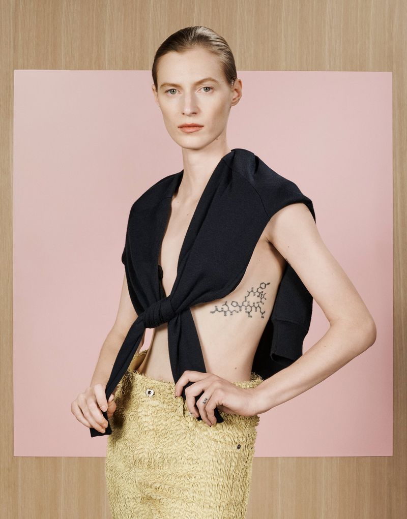 Model Julia Nobis wearing a black sweater tied over shoulders and textured yellow trousers against a pink and wood paneled backdrop