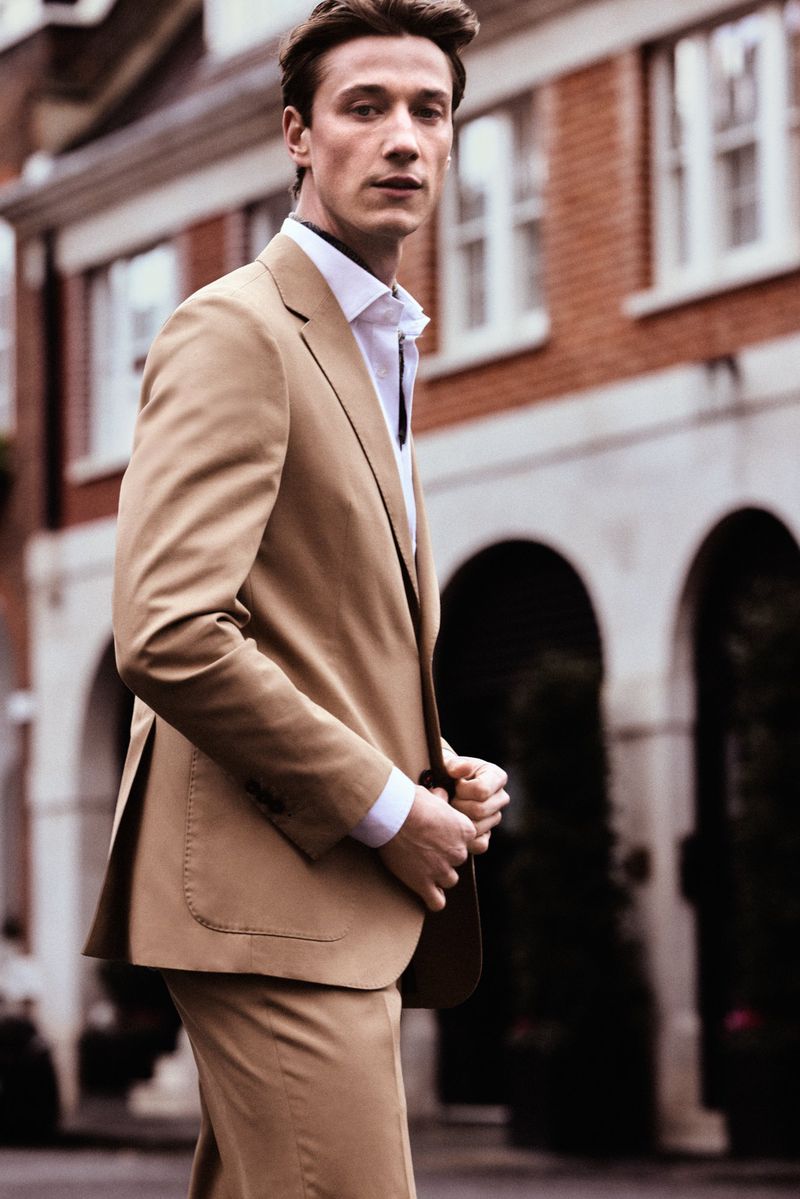 Model Harvey James in a camel suit and white shirt outdoors against a building facade for Carolina Herrera.