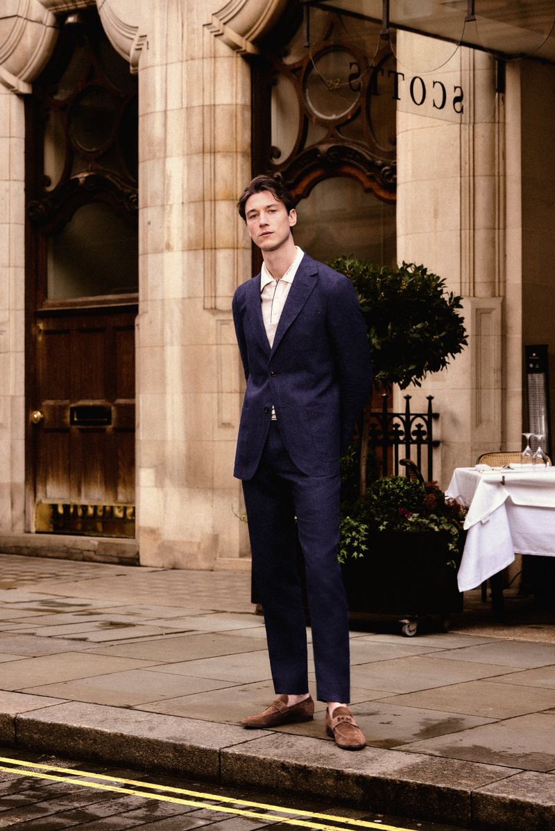 Model Harvey James in a navy blue suit standing outside a classic stone building entrance for Carolina Herrera.
