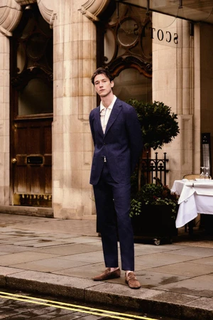 Model Harvey James in a navy blue suit standing outside a classic stone building entrance for Carolina Herrera.
