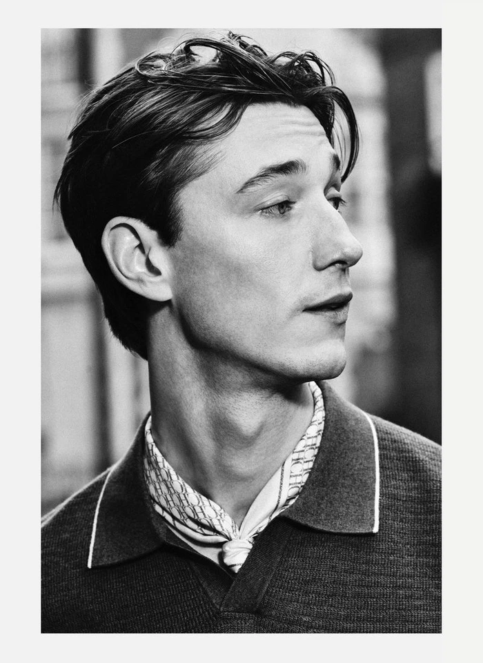 Black and white portrait of a male model wearing a patterned neckerchief and dark polo shirt looking to the side.