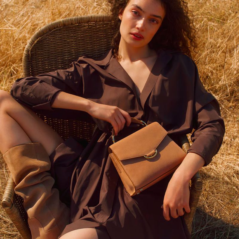 Model Sasha Kichigina in a brown shirt dress and boots holding a tan handbag while seated outdoors in a wicker chair.