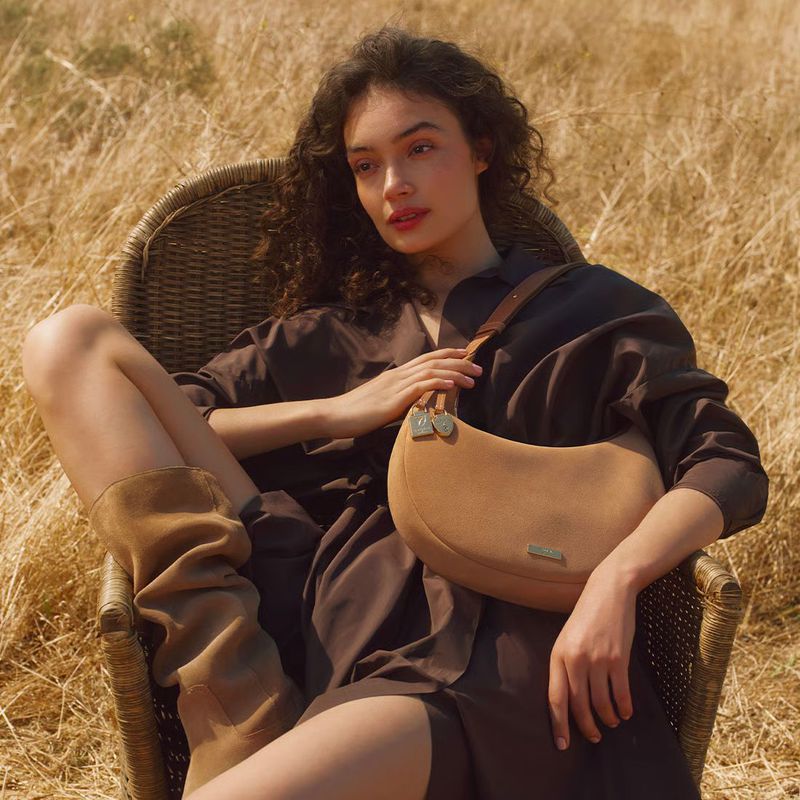 Model Sasha Kichigina in brown dress and boots holding a tan suede shoulder bag seated outdoors in a wicker chair among dry grass.