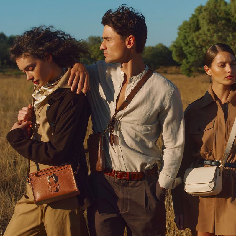 Three models wearing neutral apparel and holding leather shoulder bags outdoors in a grassy field during golden hour.