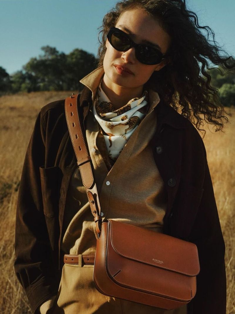 Brunette woman in a brown jacket and sunglasses, posing outdoors with a leather crossbody bag in a fashion editorial setting.