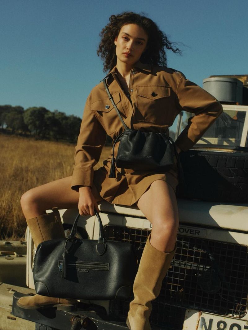 Model Sasha Kichigina in a camel safari shirt dress and tall boots posing on a vintage vehicle with black leather handbags for Aspinal Campaign S/S26.