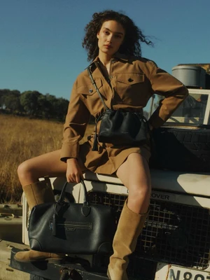 Model Sasha Kichigina in a camel safari shirt dress and tall boots posing on a vintage vehicle with black leather handbags for Aspinal Campaign S/S26.