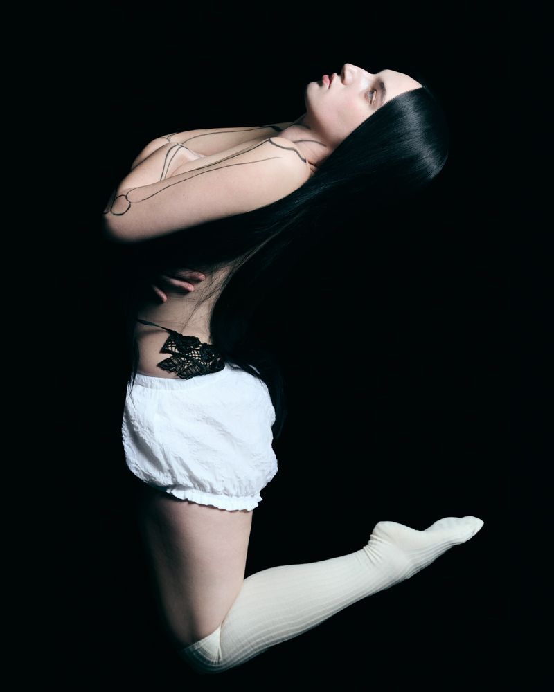 Profile view of a woman with long black hair wearing white shorts and high socks against a stark black background.
