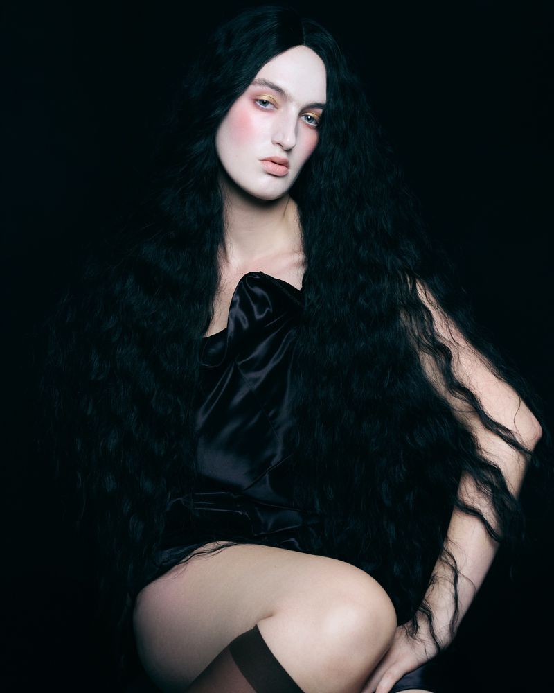 Model with long dark wavy hair and dramatic pale makeup wearing a black satin top against a dark background