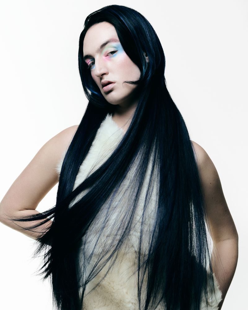 Model with long dark hair and colourful eye makeup wearing a white top against a white background for Tush Magazine