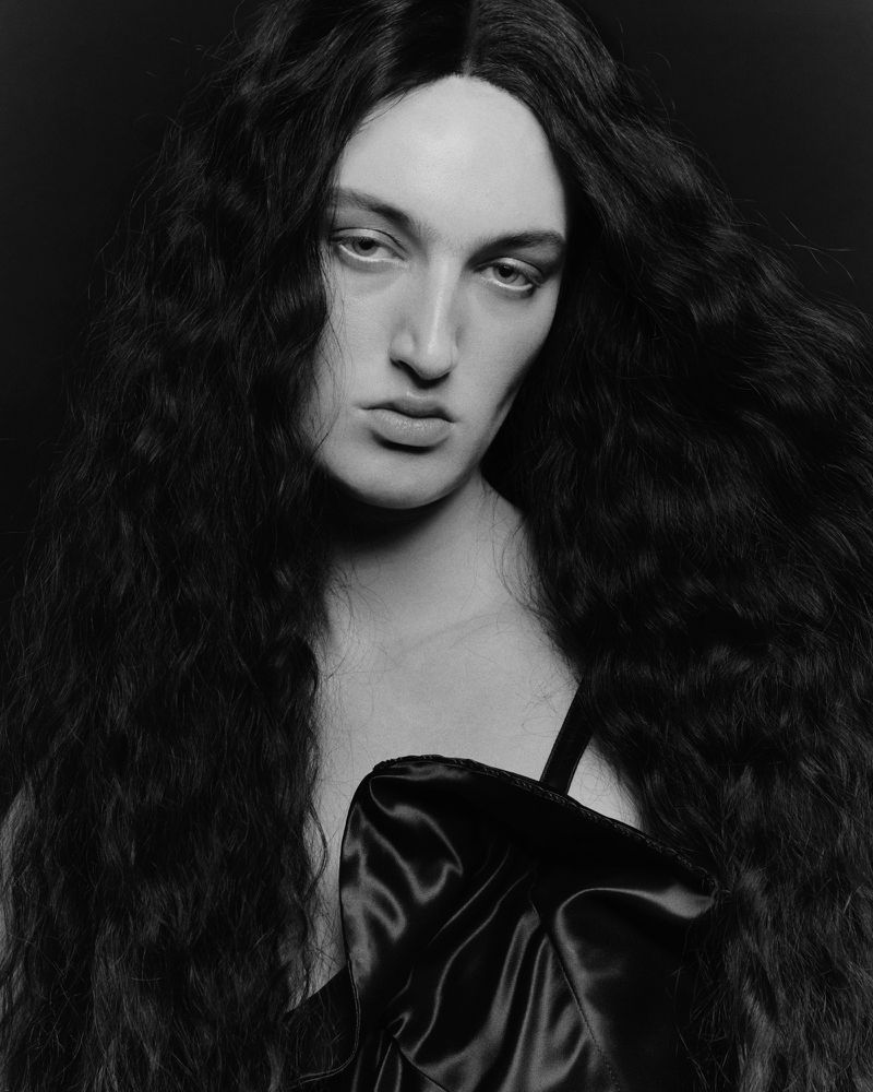 Portrait of a person with long dark wavy hair wearing a black satin top against a dark background for Tush Magazine.