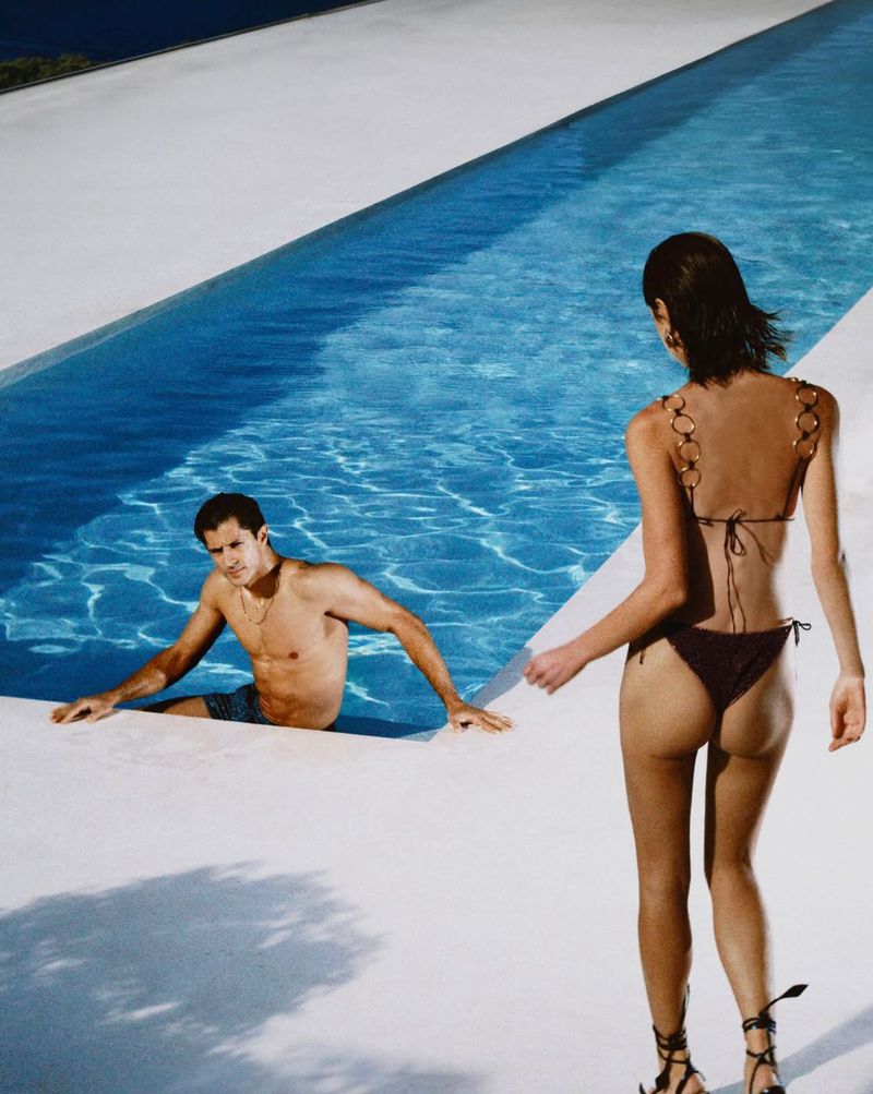 Woman in bikini walking by a swimming pool where a shirtless man is seated in the water