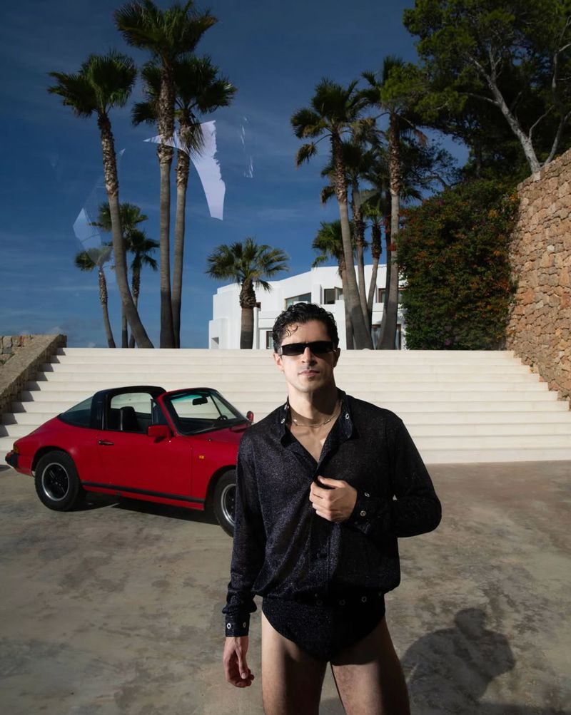 Man wearing dark sunglasses and black shirt standing outdoors with a vintage red car and white building in the background