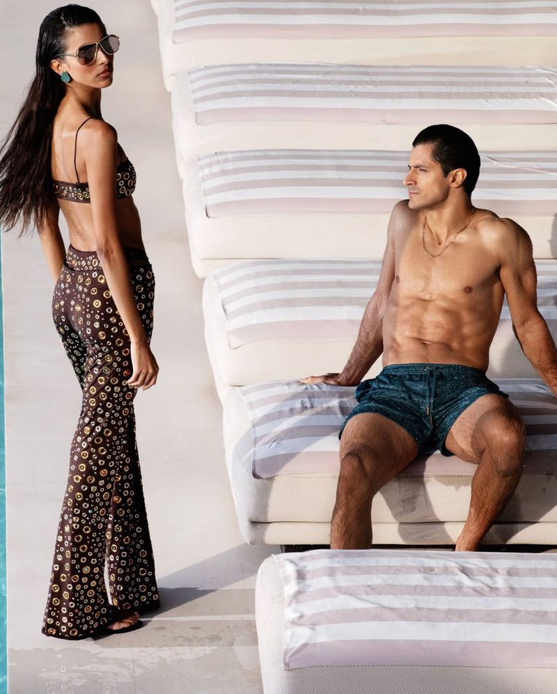 Woman in patterned two-piece outfit standing near a seated shirtless man on outdoor loungers for 70s Mania project.