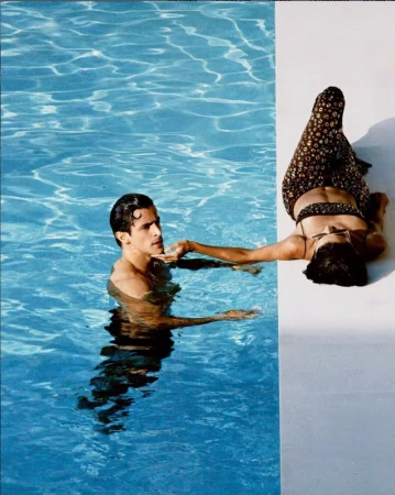 Man in swimming pool being touched on the chin by a woman lying on the white poolside ledge