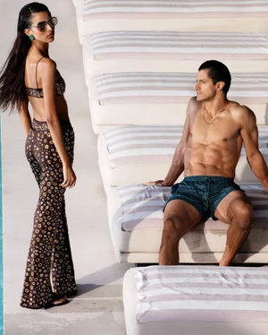 Woman in patterned two-piece outfit standing near a seated shirtless man on outdoor loungers for 70s Mania project.
