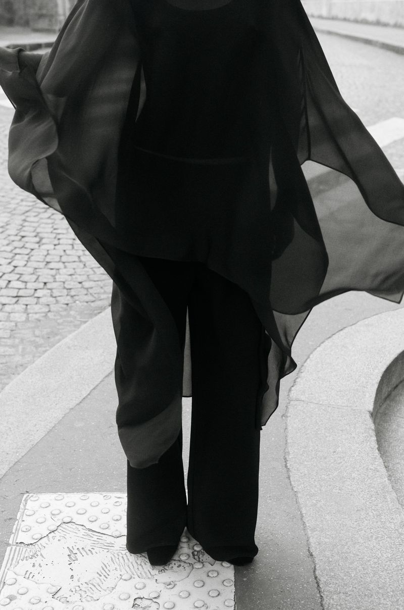 Model in flowing black sheer cape and wide trousers standing on a paved street