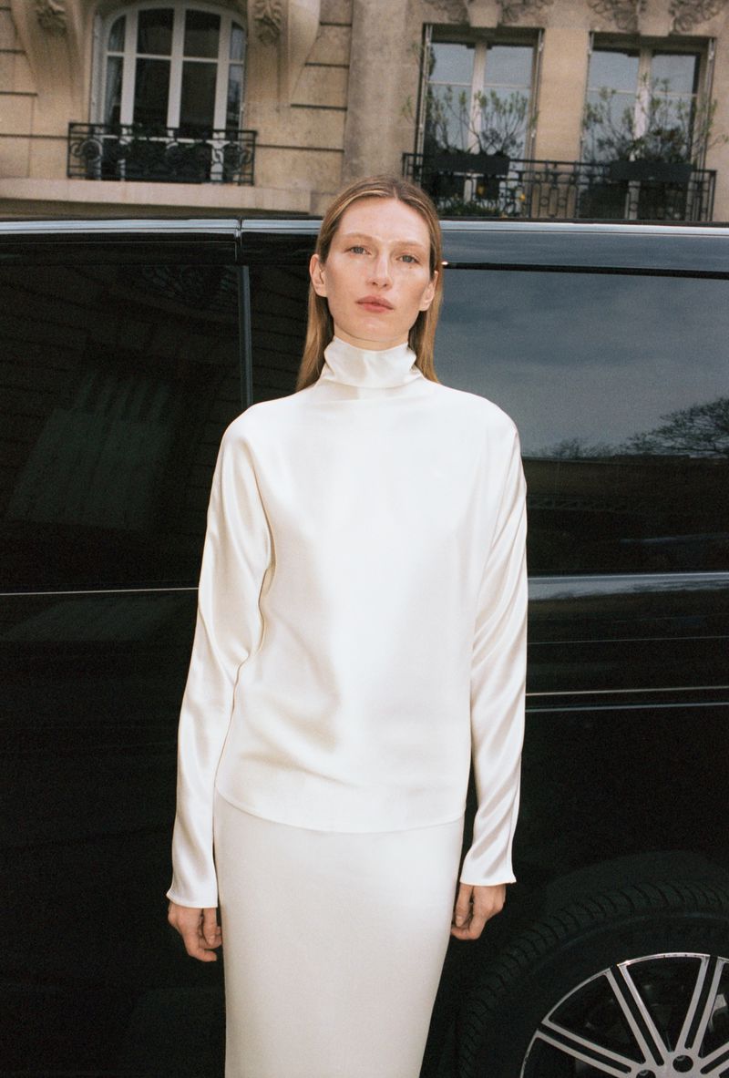 Model Liz Kennedy in a white turtleneck top and matching skirt standing in front of a black car for ALMADA S/S 2026