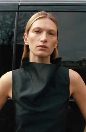 Close-up portrait of model Liz Kennedy wearing a black sleeveless top against a dark vehicle exterior for ALMADA S/S 2026.