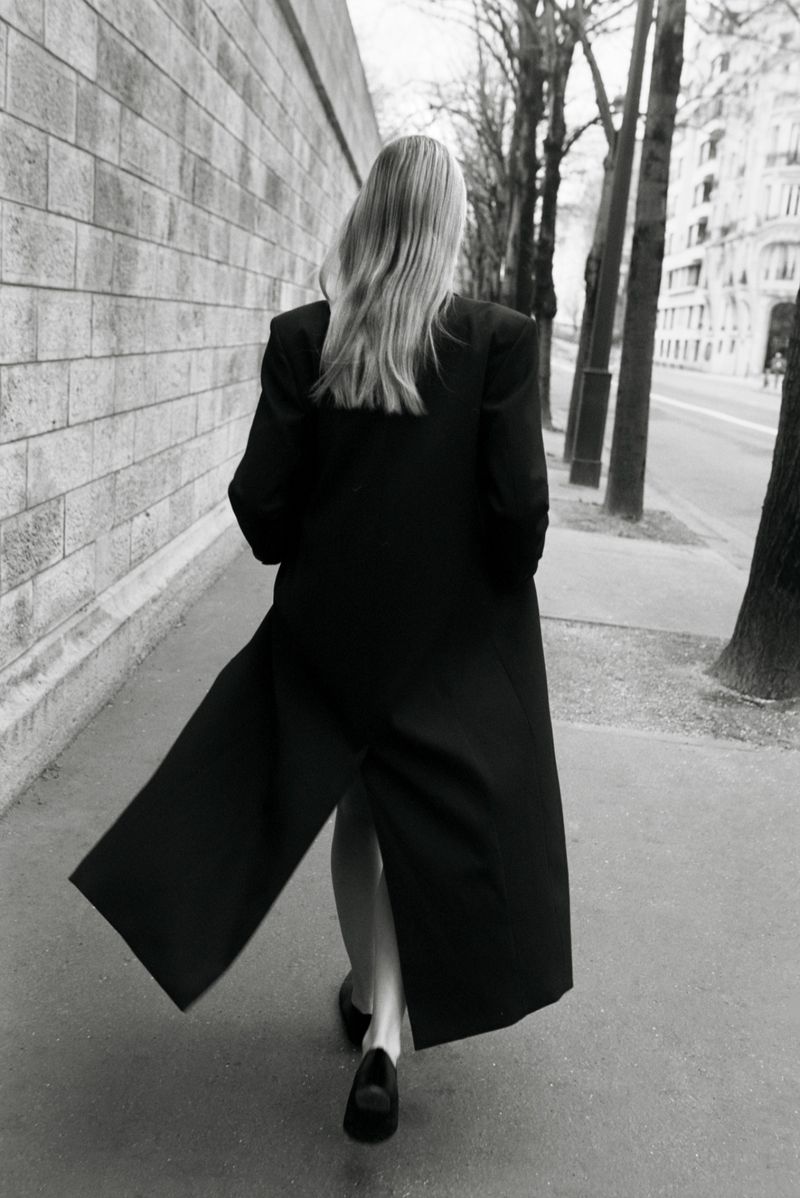 A woman with long blonde hair walks away from the camera, wearing a long black coat on a sidewalk in front of a stone wall.