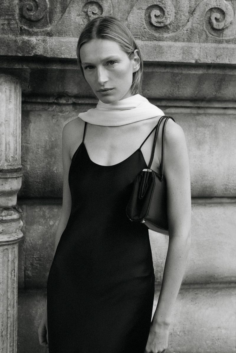 Model wearing black slip dress and light scarf holding handbag against stone architectural background