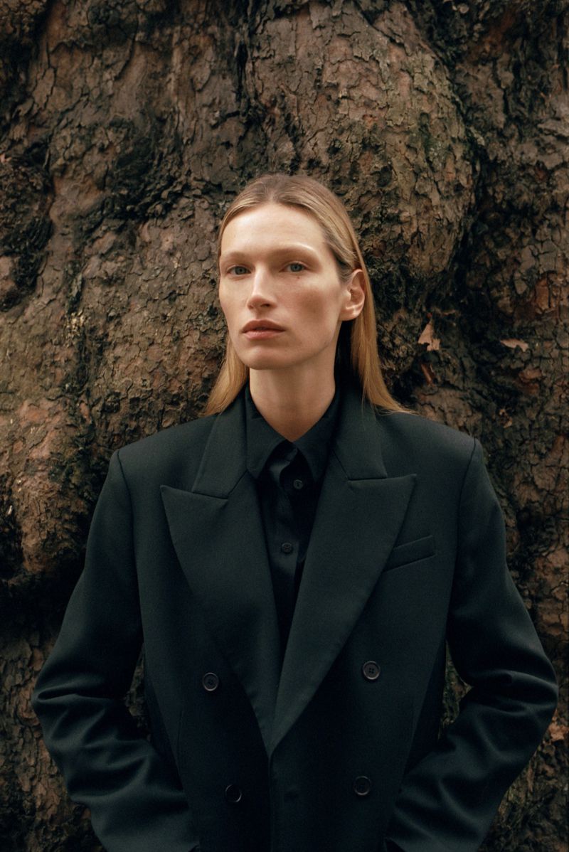 Model Liz Kennedy wearing a dark double-breasted blazer in front of a tree trunk for ALMADA S/S 2026