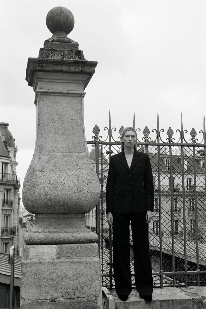 Model standing outdoors wearing a dark suit jacket and trousers near an ornate stone pillar and metal fence