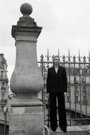 Model standing outdoors wearing a dark suit jacket and trousers near an ornate stone pillar and metal fence
