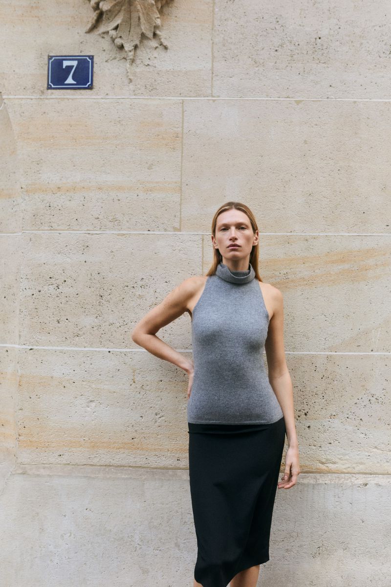 Model standing against a large stone wall wearing a grey turtleneck top and a black skirt for Almada S/S 2026