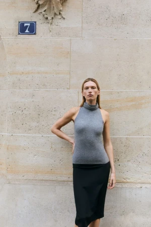 Model standing against a large stone wall wearing a grey turtleneck top and a black skirt for Almada S/S 2026