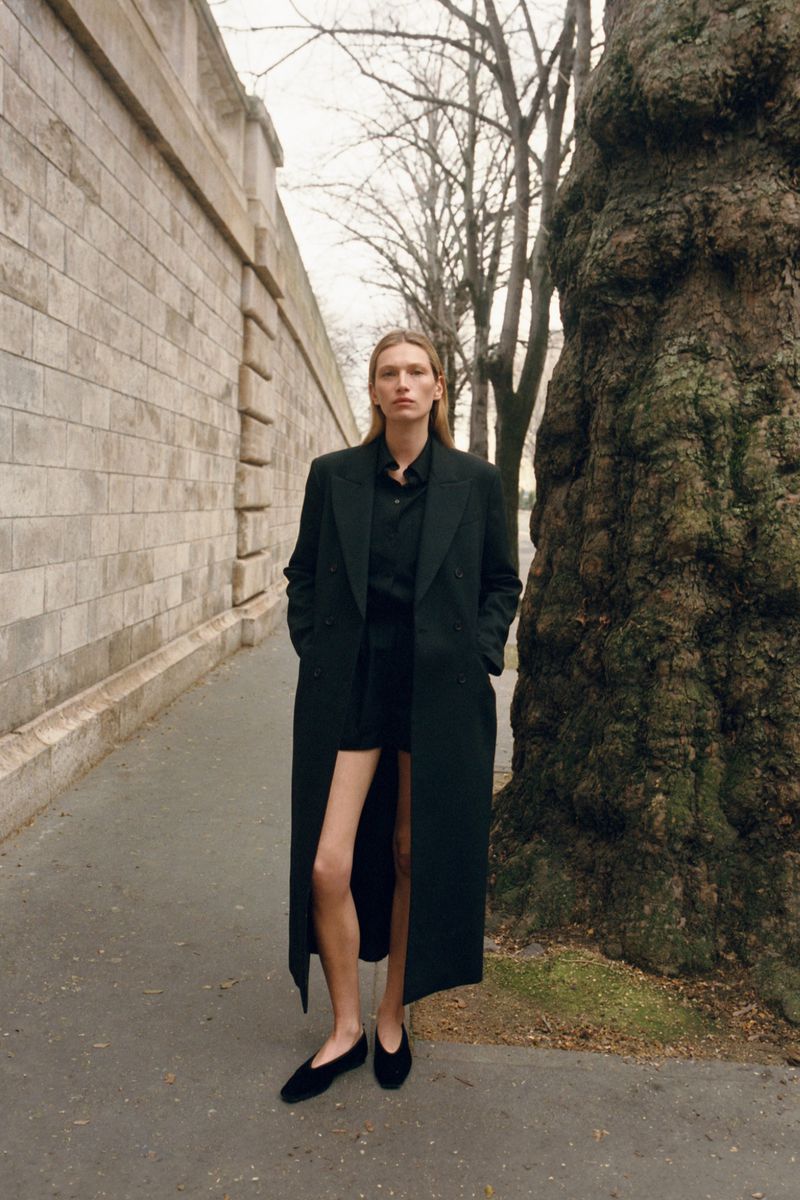 Model Liz Kennedy wearing a long black coat standing on a paved walkway beside a stone wall and tree.