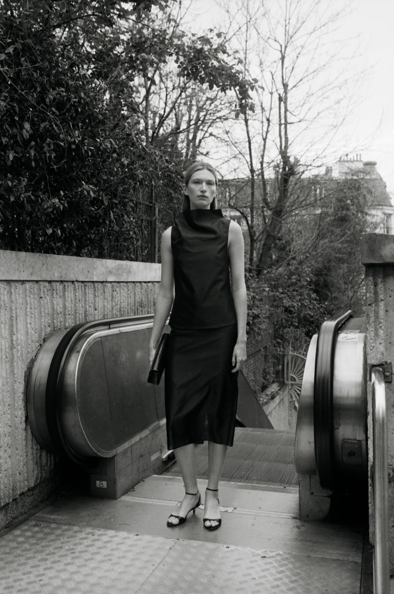 Monochrome fashion portrait of a model standing at the top of an outdoor escalator wearing a dark top and skirt holding a clutch.