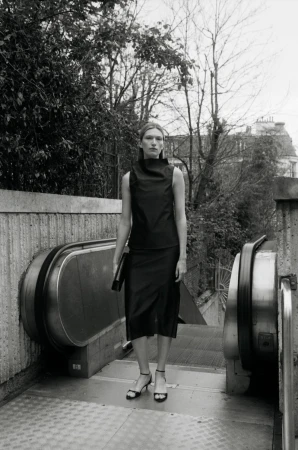 Monochrome fashion portrait of a model standing at the top of an outdoor escalator wearing a dark top and skirt holding a clutch.